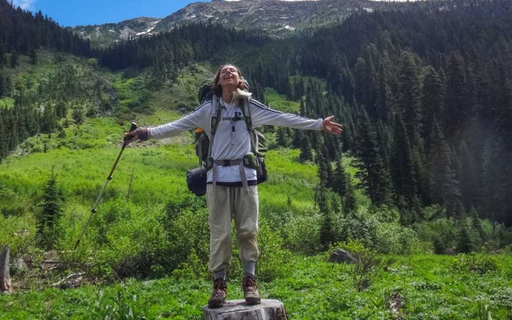 A person standing on a stump spreads their arms out wide. Behind them is a green valley.